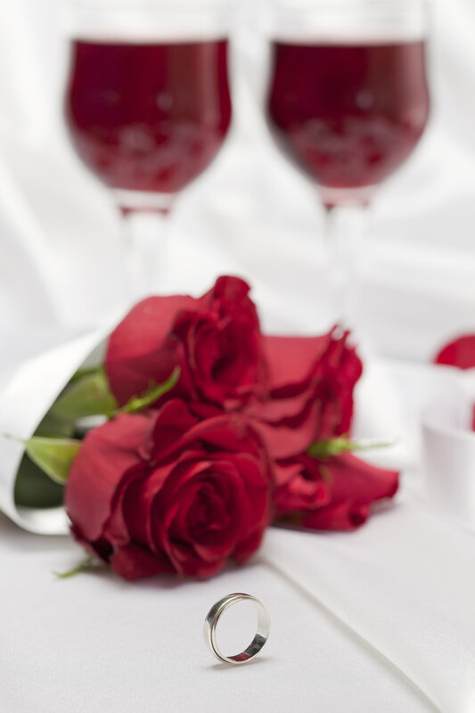 Red roses, glasses of wine and a ring on white satin background