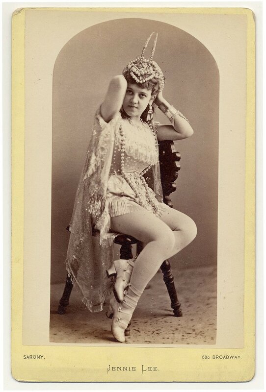 1890. Jennie Lee sitting on a chair,  holding a hair-dress on her head.